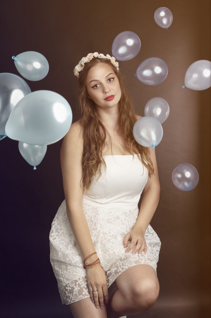 Frau mit blonden Locken in weißem Kleid, umgeben von blauen Luftballons.