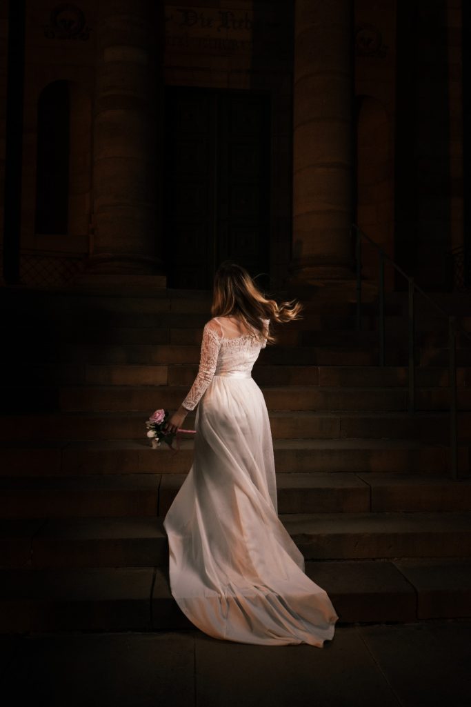 Frau in einem weißen Kleid, die eine Treppe hinaufsteigt und einen Blumenstrauß hält.