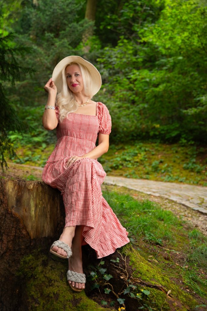 Frau in einem rosa Boutique-Kleid mit Hut sitzt auf einem Baumstamm in einem grünen Wald.
