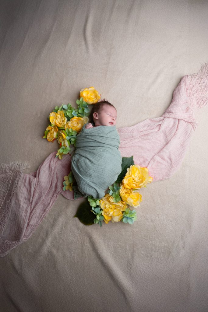 Schlafendes Baby in grüner Decke, umgeben von gelben Blumen auf hellem Hintergrund.