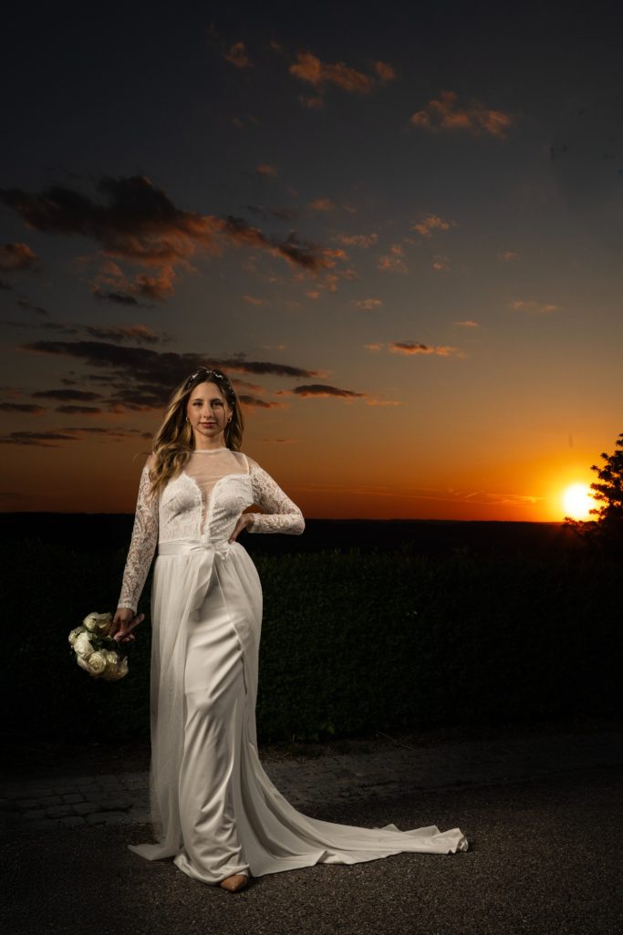 Braut in einem eleganten weißen Kleid hält einen Blumenstrauß vor einem Sonnenuntergang.
