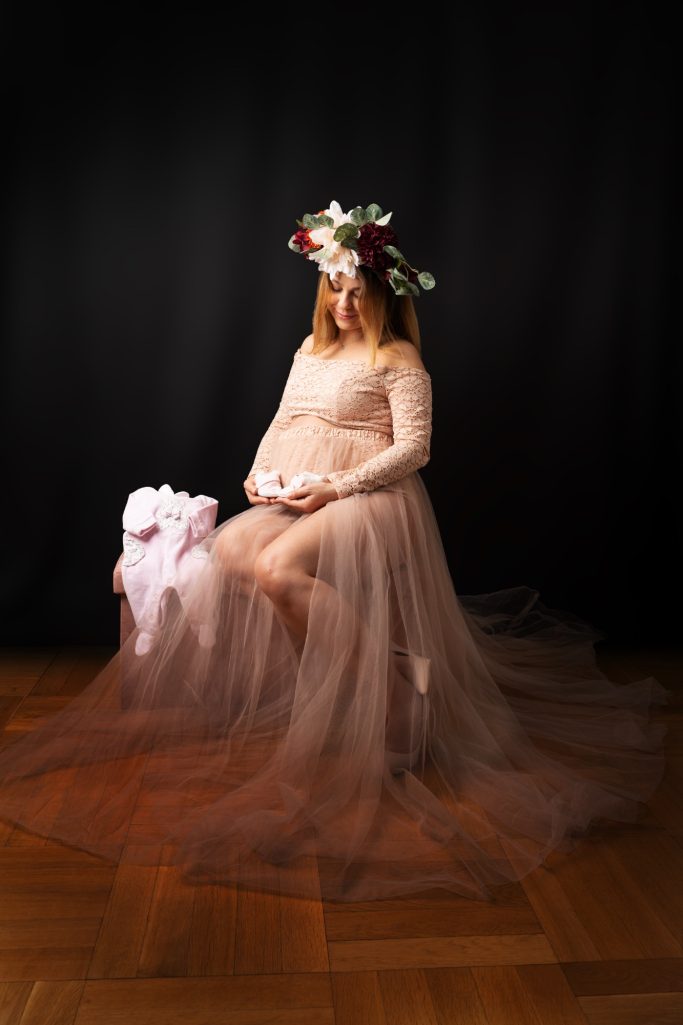 Schwangere Frau in transparentem Kleid mit Blumenkranz auf dem Kopf, sitzt auf einem Stuhl.
