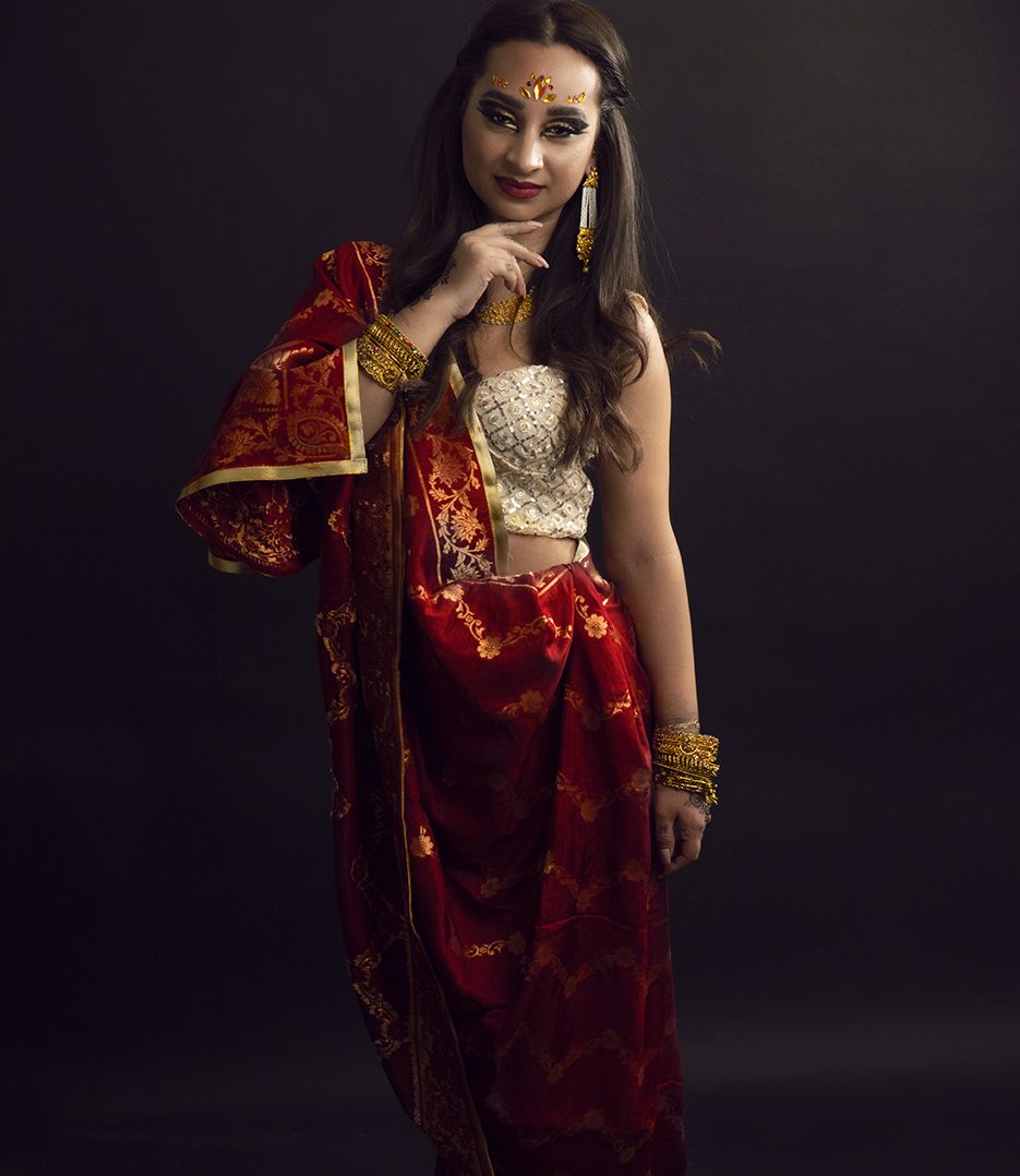 Frau in rotem Sari mit goldener Verzierung, posierend vor dunklem Hintergrund.