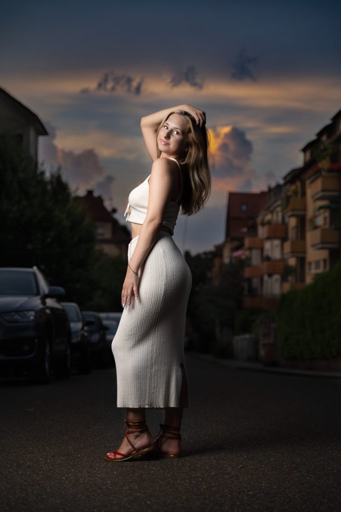 Frau in weißem Kleid posiert auf einer Straße mit Häusern und Wolken im Hintergrund.