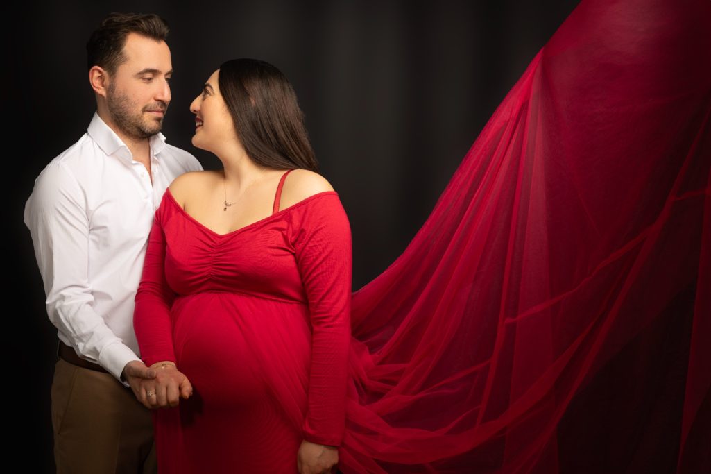 Schwangere Frau in rotem Kleid steht neben einem Mann, beide lächeln sich an.