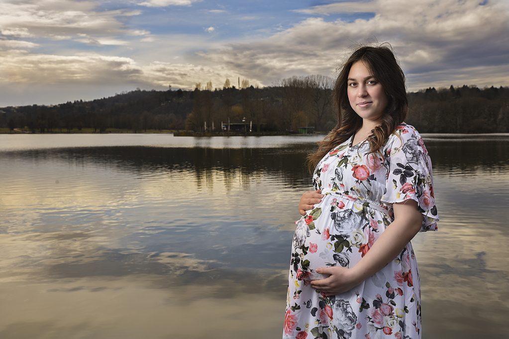 Schwangere Frau in einem blumigen Kleid steht am Wasser mit Blick auf die Landschaft.