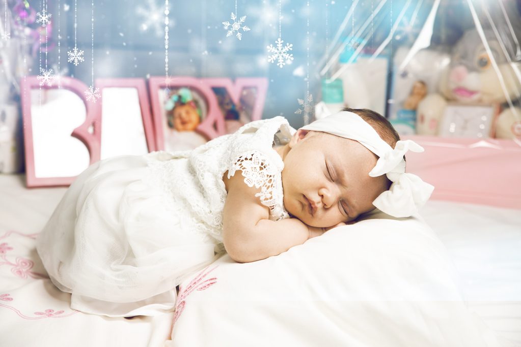 Schlafendes Baby in weißem Kleid mit großem Haarband auf einer weichen Decke.