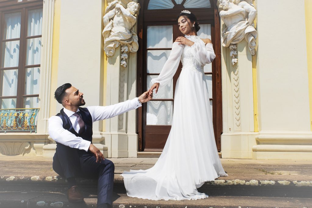 Braut in schneeweißem Kleid hält die Hand des knienden Bräutigams vor eleganter Kulisse.